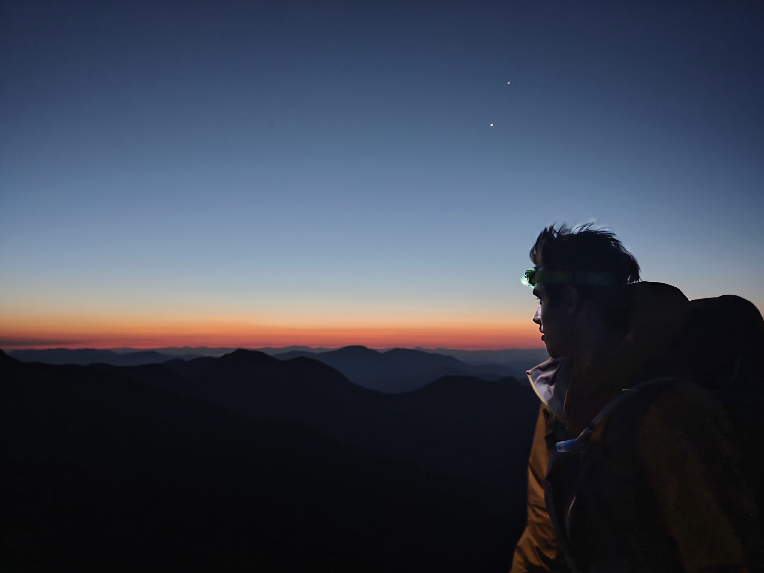 Jeune homme scrutant l'horizon montagneux à l'aube, avec un manteau et une lampe frontale.