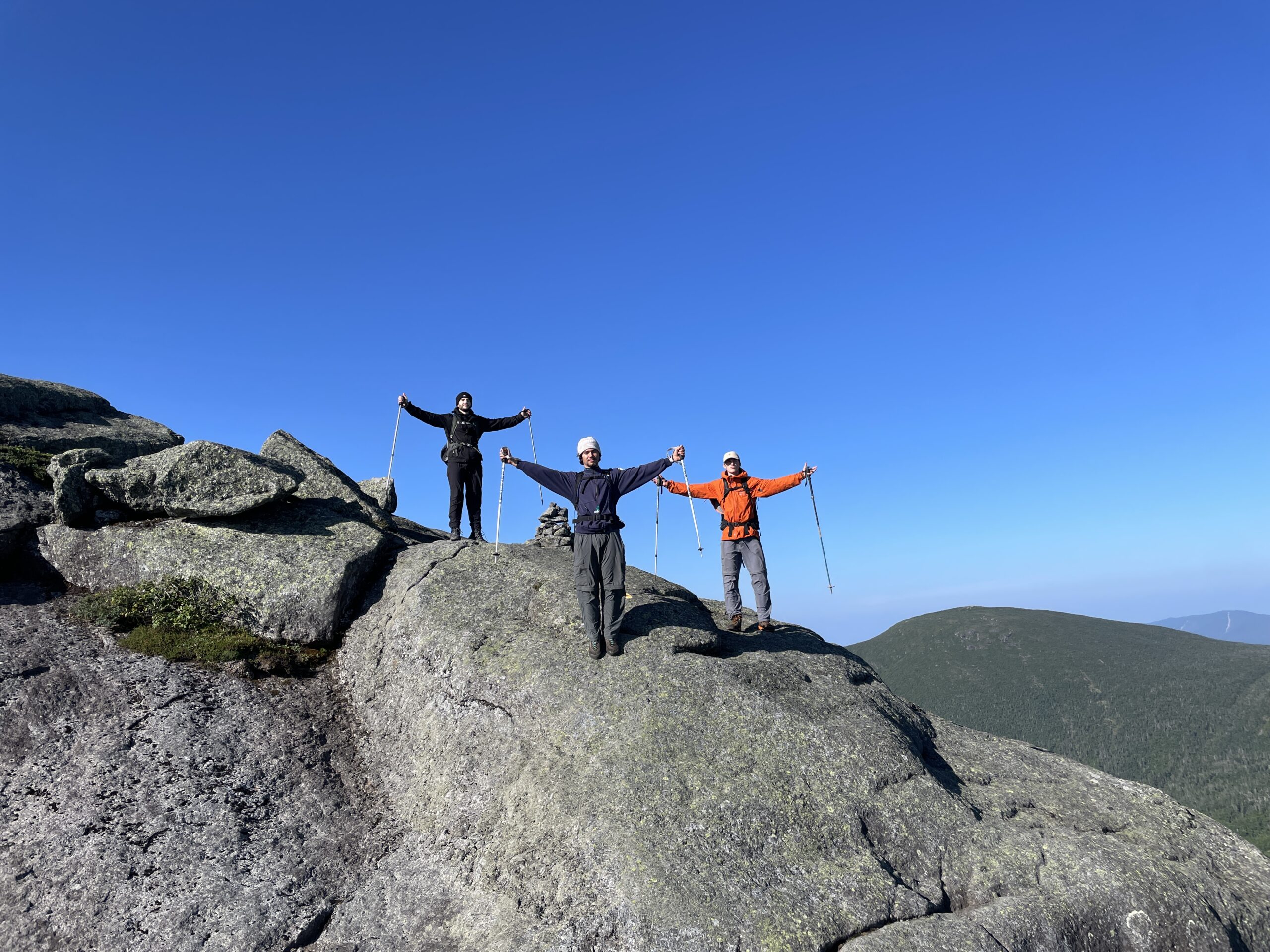 Paysage montagneux dans une lumière blanche et forte. Ciel d'un bleu éclatant. 3 jeunes hommes debouts au centre, les bvras dans les airs.